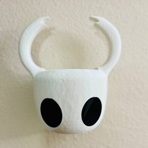 Puede incluir: Una maceta decorativa blanca, impresa en 3D, con forma de cabeza de personaje de videojuego. Presenta dos grandes aberturas negras en forma de ojos y cuernos curvos que se extienden hacia arriba. La maceta está montada en una pared de color claro.