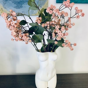 May include: White ceramic vase shaped like a human torso, holding a bouquet of pink flowers with green leaves.