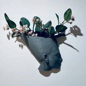 Puede incluir: Un busto de cerámica gris de una reina egipcia con un jarrón en forma de corona, lleno de flores rosas y hojas verdes.