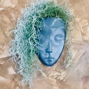 Puede incluir: Un macetero de cerámica azul con forma de cara con una larga y fluida hebra de pelo de planta de aire.