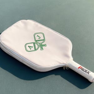 May include: A cream-coloured pickleball paddle cover with a green logo featuring the letters "A" and "J" and a pickleball design. The handle has the word "PICKK" in red. The cover is on a teal surface.