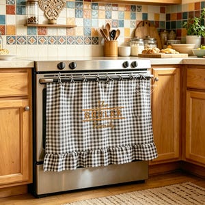 May include: A black and white gingham check stove curtain featuring a ruffled bottom edge and the embroidered text "the GOBLAR family" in a gold-toned thread, suspended from a metal rod attached to the front of a stainless steel kitchen stove.