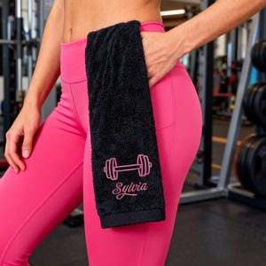 May include: Black gym towel with a pink dumbbell design and the name "Sylvia". The towel is held in the pocket of pink leggings. The person is wearing a black sports bra. The background shows gym equipment.