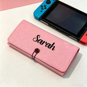 May include: A pink, rectangular case with the name "Sarah" embroidered in black script. The case has a button closure and is next to a Nintendo Switch console with blue and red controllers.