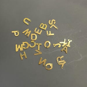 May include: A collection of gold metal letter charms, scattered on a gray surface. The letters include A, B, C, D, E, F, G, H, I, J, K, L, M, N, O, P, Q, R, S, T, U, V, W, X, Y, and Z.