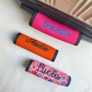 Op de afbeelding: Drie bagagehandvatwikkels in levendige kleuren. Eén is felroze met blauw geborduurde tekst "Doreen". Een andere is oranje met zwarte geborduurde tekst "Amelia". De derde is roze met een bloemenpatroon en zwarte geborduurde tekst "Lucia".