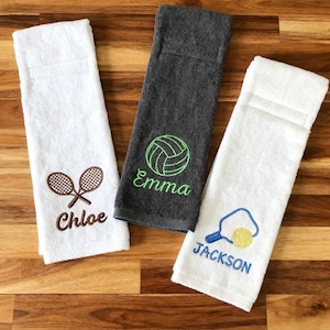 Puede incluir: Tres toallas de mano rectangulares con diseños deportivos bordados y nombres. Una toalla blanca con raquetas de tenis marrones y el nombre Chloe, una toalla gris carbón con un voleibol verde y el nombre Emma, y una toalla blanca con una pala de pickleball azul y amarilla y el nombre Jackson.