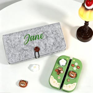 May include: A gray felt case with the name "Jane" in green script, a brown button closure, and a green Switch controller with bear and flower designs. Also, two small decorative items.