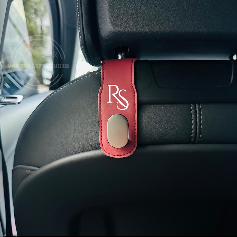 May include: A red leather car seat headrest hook with the white letters "RS" and a silver-colored magnetic clip. The hook is designed to hang items in a vehicle. The background shows a car interior.