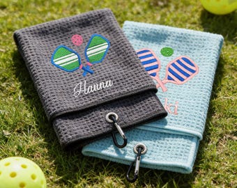 May include: Two waffle-weave pickleball towels, one gray and one light blue, embroidered with pickleball paddles and balls. The gray towel has the name "Hanna" embroidered in white. Both towels have a black carabiner clip attached.