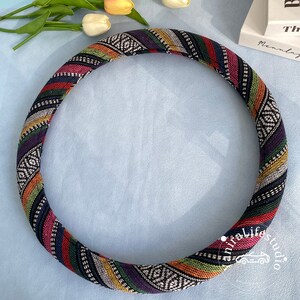 May include: A colorful, striped steering wheel cover with a black background and white geometric patterns. The cover is made of fabric and has a thick, comfortable feel.