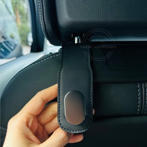 May include: A black car seat headrest hook with a silver-colored oval accent. The hook is made of a smooth, leather-like material and is designed to attach to the headrest posts. The stitching is white.