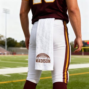May include: A white athletic towel hanging from the waistband of white football pants with maroon and gold side stripes. The towel features an embroidered brown football graphic above the text "MADISON" in brown lettering.