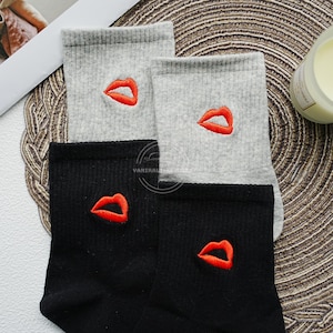 May include: A pair of gray and a pair of black socks. Each sock features a red embroidered lip design. The socks are ribbed and appear to be made of a soft material. The socks are folded and displayed together.