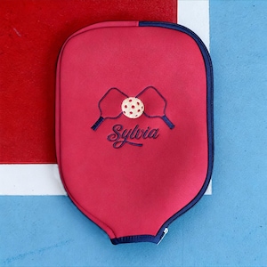 Op de afbeelding: Een rode pickleball paddle cover met een marineblauwe rits en rand. De hoes heeft een geborduurd ontwerp van twee pickleball paddles en een bal, met de naam "Sylvia" in cursief.