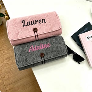Puede incluir: Dos estuches de fieltro personalizados, uno rosa y otro gris, cada uno con un nombre bordado en la parte delantera. El estuche rosa dice "Lauren" en negro, y el estuche gris dice "Adaline" en rosa. Ambos tienen un cierre de botón.
