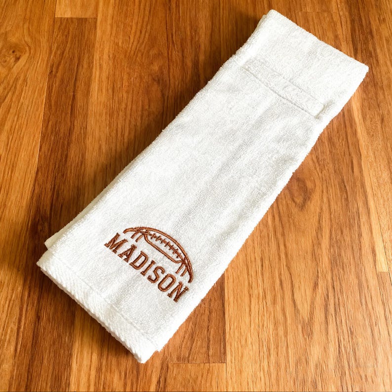 May include: A white hand towel featuring brown embroidery of an American football icon positioned above the name MADISON in capital letters. The towel has a soft, textured fabric surface and is folded into a rectangular shape.