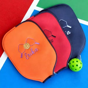 Op de afbeelding: Drie pickleball paddle covers in oranje, rood en marineblauw, elk met een ritssluiting. De oranje cover heeft een paars geborduurd ontwerp met een pickleball en de naam "Erika". Een groene pickleball ligt ernaast.