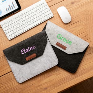 Könnte beinhalten: Zwei graue und schwarze Laptop-Hüllen aus Filz mit in Pink und Grün gestickten Namen. Jede Hülle hat einen braunen Lederakzent und einen Klappenverschluss. Eine weiße Tastatur, Maus und ein Tablet befinden sich im Hintergrund.