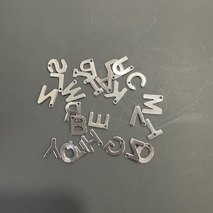 Peut inclure: Un ensemble de breloques en m&eacute;tal argent&eacute; en forme de lettres, &eacute;parpill&eacute;es sur une surface grise. Les lettres sont toutes en majuscules et comprennent A, B, C, D, E, F, G, H, I, J, K, L, M, N, O, P, Q, R, S, T, U, V, W, X, Y et Z.