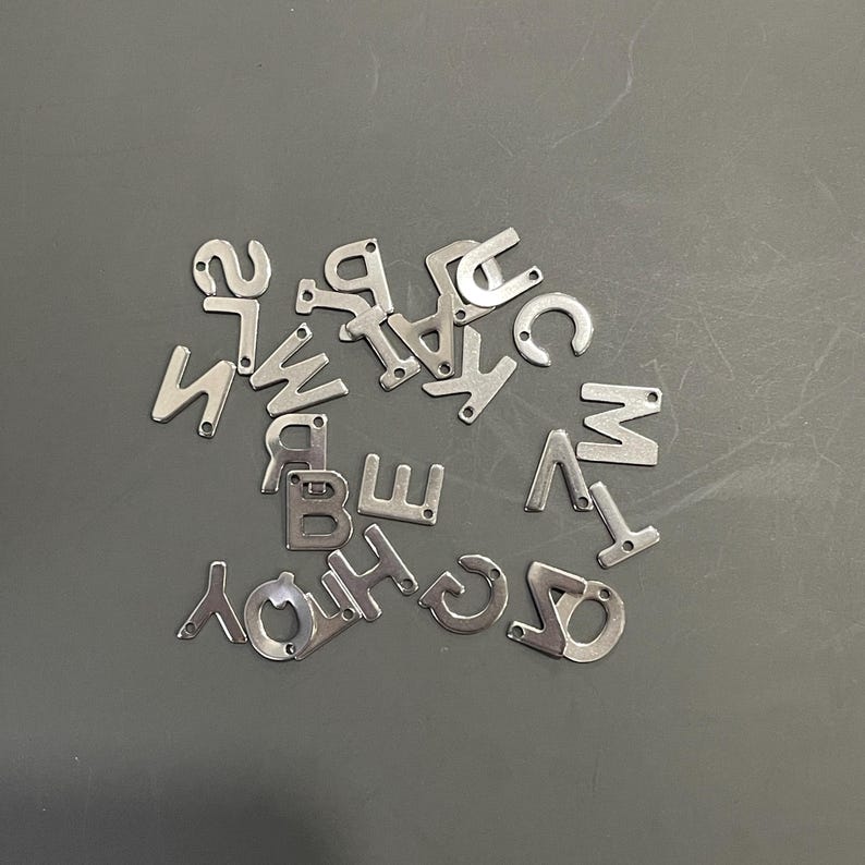 Peut inclure: Un ensemble de breloques en m&eacute;tal argent&eacute; en forme de lettres, &eacute;parpill&eacute;es sur une surface grise. Les lettres sont toutes en majuscules et comprennent A, B, C, D, E, F, G, H, I, J, K, L, M, N, O, P, Q, R, S, T, U, V, W, X, Y et Z.