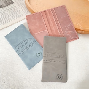 May include: A collection of car registration and insurance holders in various colors. The holders are light blue, pink, and gray, with a car outline and the words "REGISTRATION & INSURANCE" printed on them. Each holder has a different initial in a wreath.