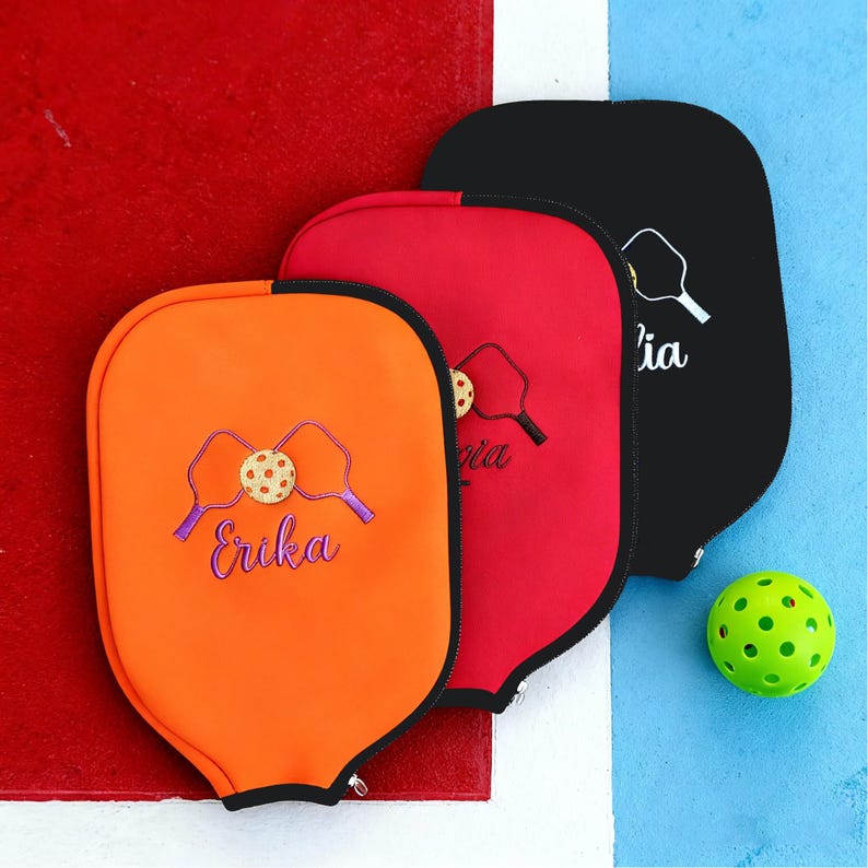Op de afbeelding: Drie pickleball paddle covers in oranje, rood en zwart, elk met een ritssluiting. De oranje cover heeft een pickleball- en paddle-ontwerp met de naam "Erika". Een groene pickleball ligt in de buurt.