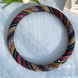 May include: A colorful, patterned steering wheel cover with a black background and stripes of red, yellow, green, and white. The cover has a woven texture and is decorated with small metal studs.