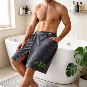 May include: A charcoal gray terry cloth towel wrap featuring a front pocket, a snap button closure at the waist, and the embroidered green monogram letters ASM on the lower right side. The towel wrap is made of a soft, absorbent fabric.