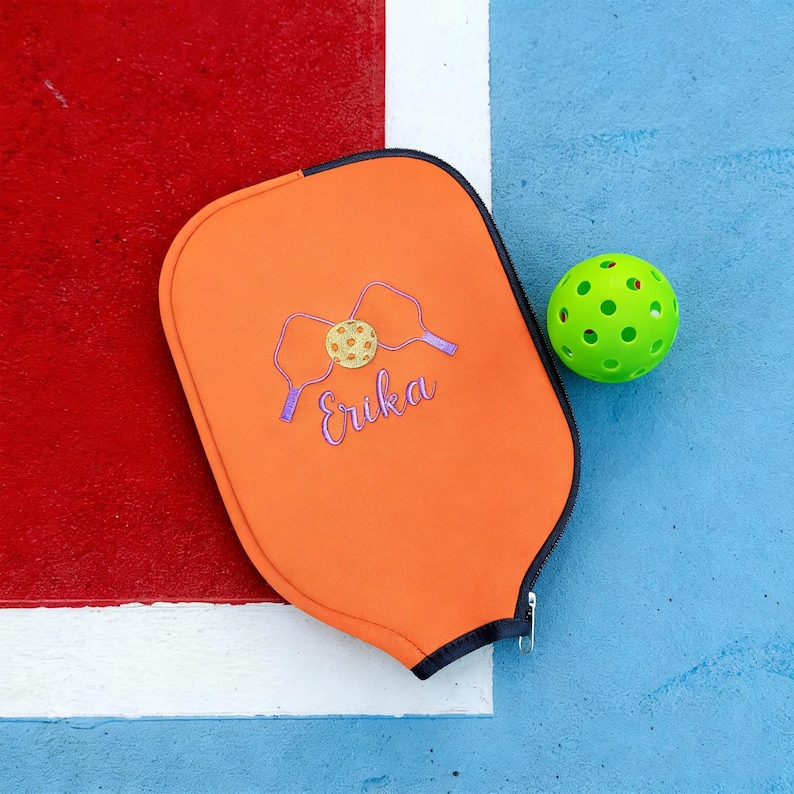 Op de afbeelding: Een oranje pickleball paddle cover met een rits, geborduurd met een pickleball- en paddle-ontwerp, en de naam "Erika" in paarse letters. Een groene pickleball ligt naast de cover op een blauw oppervlak.