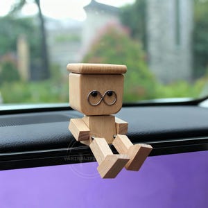 以下が含まれることがあります： 正方形の頭と体、関節式の腕と脚を持つ小さなロボットの形をした木製の置物。 ロボットは銀色のワイヤーの目と小さな木の帽子をしています。 黒い表面に座っています。