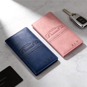 May include: Two leather car registration and insurance holders, one navy blue and one pink. Each features a car outline and the text "REGISTRATION & INSURANCE". The navy holder has "N.B.P" and the pink holder has "R.L.Y" engraved.