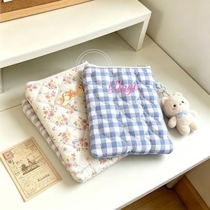 May include: Two quilted pouches, one with a floral pattern and the other with a blue and white gingham check. The floral pouch has the name "Blue" embroidered on it, and the gingham pouch has the name "Hugo". A small teddy bear keychain is attached to the gingham pouch.