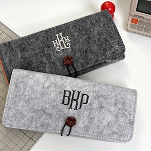 May include: Two felt cases, one dark gray and one light gray, each with a monogram. The dark gray case has a white monogram and an orange interior. The light gray case has a black monogram. Both cases have a button closure.