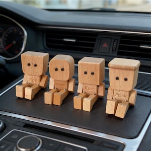 Könnte beinhalten: Vier Holzfiguren mit beweglichen Armen und Beinen, jede mit einem eingravierten Namen: Eric, Diana, Rose und Frank. Die Figuren haben schwarze Punktaugen und sitzen auf einem Armaturenbrett im Auto.