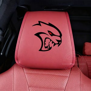 Könnte beinhalten: Rote Auto-Kopfstütze mit einer schwarz-roten Grafik eines knurrenden katzenartigen Wesens. Die Kopfstütze besteht aus einem glatten, lederartigen Material und ist Teil des Innenraums eines Autos.