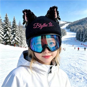 May include: A black knit beanie featuring plush cat ears with pink inner accents and small black bows. The front displays the pink embroidered text Blythe with a skier icon. The beanie is paired with large ski goggles with a reflective blue lens.