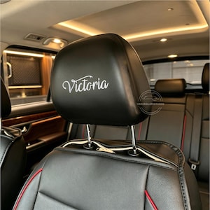 May include: Black car seat headrest with the name "Victoria" in white script. The seat is black leather with red stitching accents. The interior of the car is visible in the background.