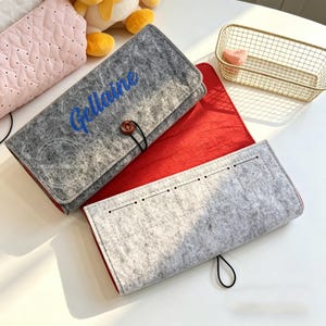 May include: Gray felt case with the name "Gellaine" embroidered in blue. The case has a button closure and a black elastic loop. A red felt lining is visible. A second gray felt case is also present.