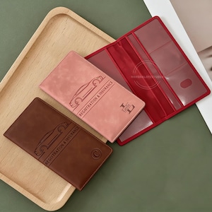 May include: Three leather registration and insurance holders in brown and pink, with a red interior. The brown holder has a car outline and the text "REGISTRATION & INSURANCE". The pink holder has the same text and a name. The red holder is open.