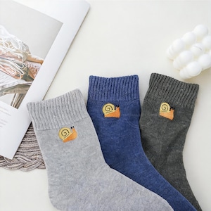 May include: Three pairs of socks in gray, blue, and charcoal gray. Each sock features an embroidered orange snail design on the cuff. The socks are made of a soft, knitted material, perfect for casual wear.