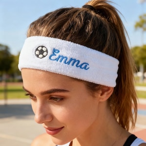 Op de afbeelding: Witte badstof hoofdband met de naam "Emma" in blauw geborduurd, samen met een voetbalafbeelding. De hoofdband wordt gedragen door een persoon met bruin haar dat in een paardenstaart is vastgebonden.