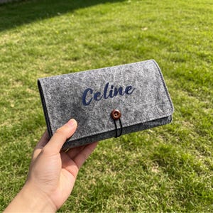 May include: Gray felt wallet with the name "Celine" embroidered in blue script. The wallet has a button closure and a black elastic loop. The background is green grass.