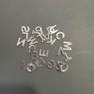 May include: A collection of silver metal letter charms, scattered on a gray surface. The letters are all uppercase and include A, B, C, D, E, F, G, H, I, J, K, L, M, N, O, P, Q, R, S, T, U, V, W, X, Y, and Z.