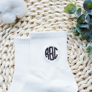 Puede incluir: Calcetines tobilleros blancos con textura acanalada, con un monograma bordado en gris oscuro de las letras "RBC" en un diseño circular. Los calcetines se exhiben sobre una superficie tejida con vegetación y una bola de algodón.