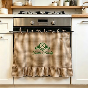 May include: A beige and white gingham oven curtain featuring a ruffled bottom edge and a green embroidered monogram with the letter S inside a decorative frame above the text Smiths Family, hung on a black rod with fabric ties across the front.