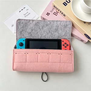 May include: A pink felt case with a gray felt interior holds a black handheld gaming console with blue and red controllers. The case has a quilted design on the front and a black loop closure. The text "Sweet of Jeans" is visible on a small card.