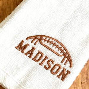 May include: A white terry cloth towel featuring brown embroidery of an American football positioned above the name MADISON in capital letters. The embroidery is stitched directly onto the fabric surface of the white towel.