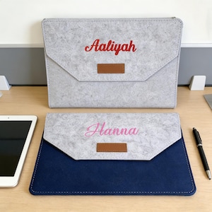 May include: Two personalized felt tablet cases. One is light gray with the name "Aaliyah" in red script, and the other is navy blue with the name "Hanna" in pink script. Both have a brown leatherette accent.