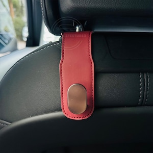 May include: A red leather car seat headrest hook with a metallic oval hook. The hook is attached to a black car seat. The stitching on the hook is red.